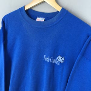 May include: A royal blue crew neck sweatshirt with the words "North Carolina" and a logo printed on the front. The sweatshirt is made of a soft material and has a classic fit.