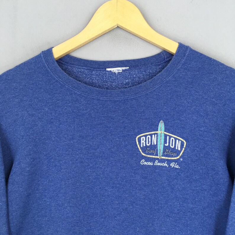 May include: A blue crewneck sweatshirt featuring a graphic on the chest with the text RON JON Surf Shop Cocoa Beach, Fla. and an illustration of a surfboard. The garment has a ribbed neckline and is displayed on a wooden hanger.