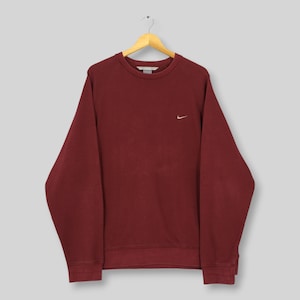 Może przedstawiać: Bordowa bluza z małym, białym logo Nike na piersi. Top z długim rękawem wisi na drewnianym wieszaku. Bluza ma okrągły dekolt i luźny kr&oacute;j.