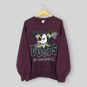 May include: A maroon sweatshirt with the Mighty Ducks of Anaheim logo. The logo features a hockey mask, crossed hockey sticks, and a hockey puck. The text "MIGHTY DUCKS" and "DUCKS OF ANAHEIM" are also displayed.