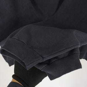 Op de afbeelding: Close-up van een marineblauwe sweatshirtmouw. De manchet is geribbeld en donkerblauw. De stof van de mouw is effen marineblauw. De mouw wordt vastgehouden door een gehandschoende hand.