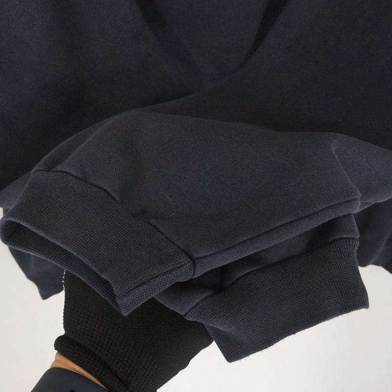 Op de afbeelding: Close-up van een marineblauwe sweatshirtmouw. De manchet is geribbeld en donkerblauw. De stof van de mouw is effen marineblauw. De mouw wordt vastgehouden door een gehandschoende hand.