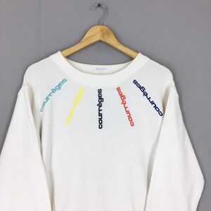 May include: White sweatshirt with the word "courr&egrave;ges" embroidered in blue, red, yellow, and green. The text is arranged in a radial pattern across the chest. The sweatshirt is hanging on a wooden hanger.