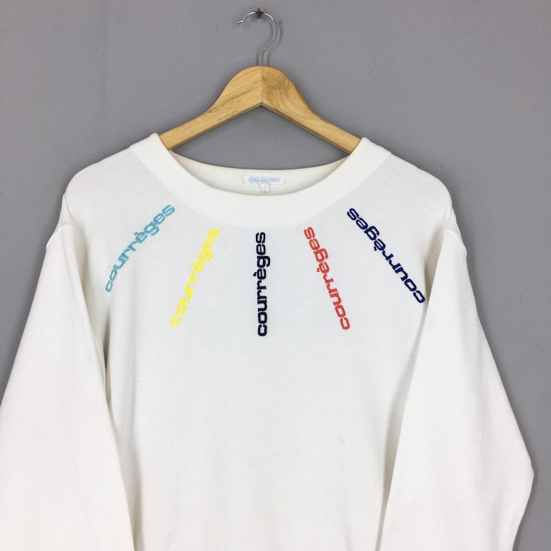 May include: White sweatshirt with the word "courr&egrave;ges" embroidered in blue, red, yellow, and green. The text is arranged in a radial pattern across the chest. The sweatshirt is hanging on a wooden hanger.