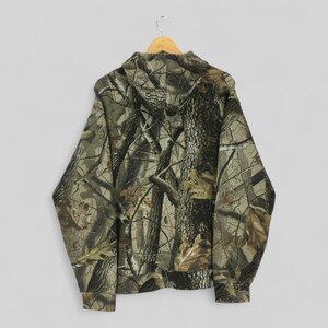 Peut inclure: Un sweat &agrave; capuche camouflage, pr&eacute;sentant un motif forestier en nuances de marron, vert et beige. Le dos du sweat &agrave; capuche est visible, suspendu &agrave; un cintre en bois. Le motif est con&ccedil;u pour se fondre dans un environnement forestier.
