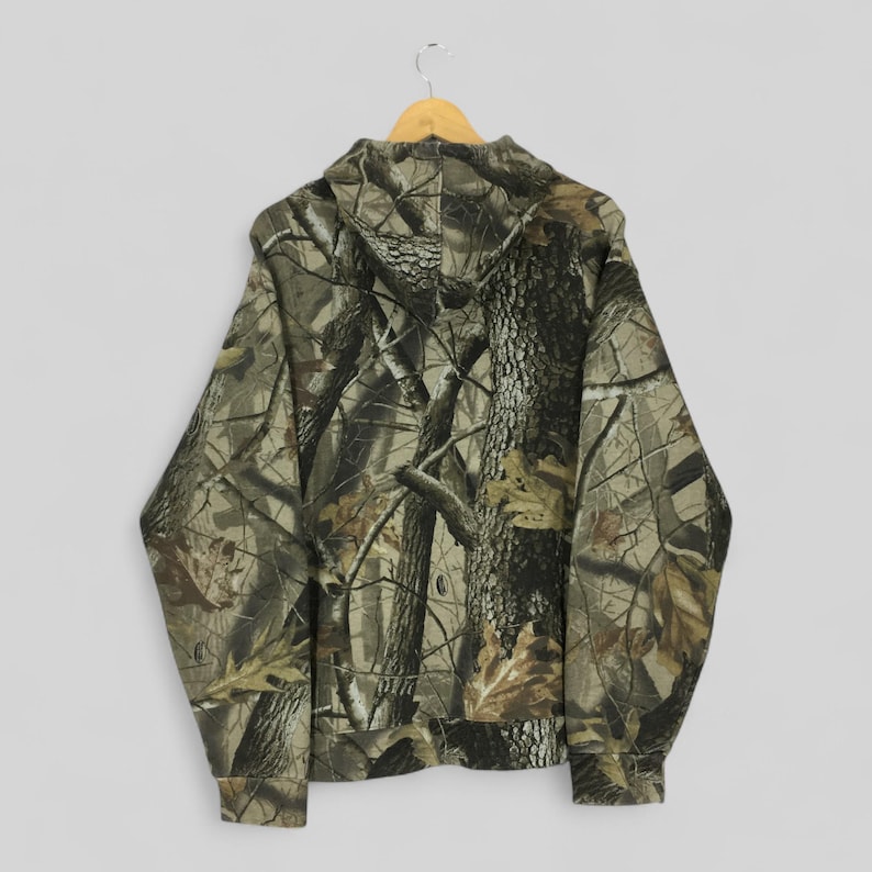 Peut inclure: Un sweat &agrave; capuche camouflage, pr&eacute;sentant un motif forestier en nuances de marron, vert et beige. Le dos du sweat &agrave; capuche est visible, suspendu &agrave; un cintre en bois. Le motif est con&ccedil;u pour se fondre dans un environnement forestier.