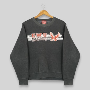 Pode incluir: Camisola cinza escuro EWU Eagles com um gráfico branco e vermelho desgastado. O design inclui o texto "EWU EAGLES" e um logotipo de águia vermelha. A camisola tem gola redonda e mangas compridas.
