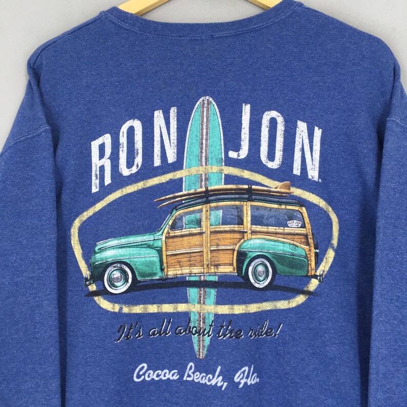 May include: A blue long-sleeve sweatshirt featuring a graphic print of a green woody station wagon with a surfboard on top. The text reads RON JON, It's all about the ride!, and Cocoa Beach, Fla. The design is centered within a yellow oval.