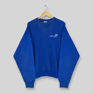 May include: A royal blue sweatshirt with the words "North Carolina" and a logo printed in light blue. The sweatshirt has a crew neck and long sleeves with ribbed cuffs and hem. The garment is hanging on a wooden hanger.