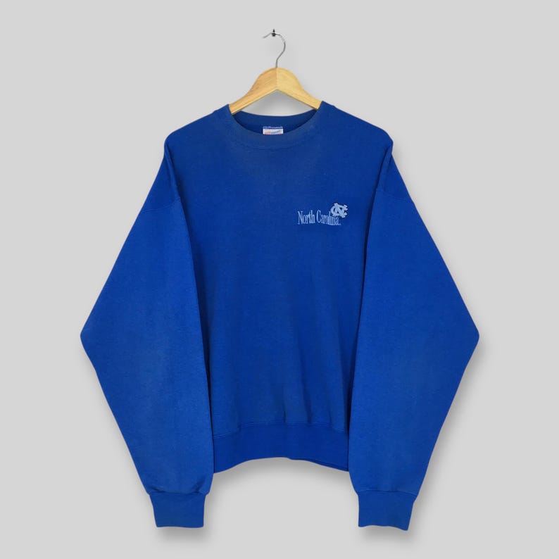 May include: A royal blue sweatshirt with the words "North Carolina" and a logo printed in light blue. The sweatshirt has a crew neck and long sleeves with ribbed cuffs and hem. The garment is hanging on a wooden hanger.