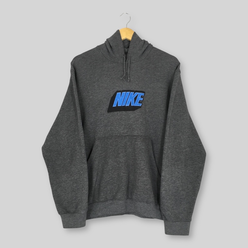Może przedstawiać: Ciemnoszara bluza z kapturem ze sznurkiem i przednią kieszenią. Słowo "NIKE" jest wyświetlane niebieskimi literami blokowymi z czarnym obrysem na piersi. Bluza z kapturem wisi na drewnianym wieszaku.