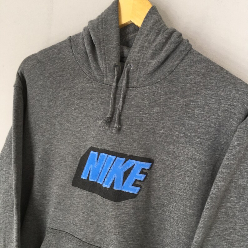Może przedstawiać: Szara bluza z kapturem z logo Nike w kolorze niebieskim i czarnym. Bluza ma kaptur ze sznurkiem i przednią kieszeń. Logo Nike znajduje się na piersi.
