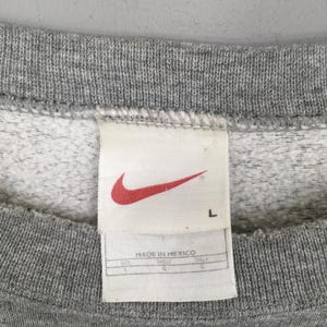 May include: Close-up of a gray sweatshirt with a white tag. The tag features a red Nike swoosh logo and the text "MADE IN MEXICO". The size is listed as L.