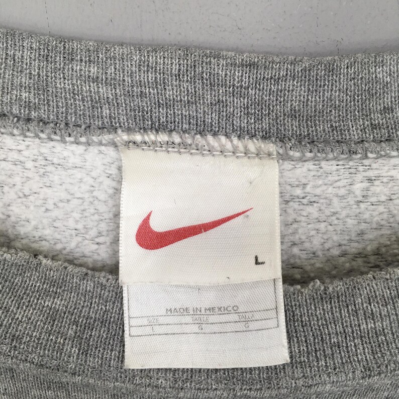 May include: Close-up of a gray sweatshirt with a white tag. The tag features a red Nike swoosh logo and the text "MADE IN MEXICO". The size is listed as L.