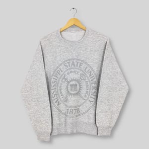 Puede incluir: Una sudadera gris jaspeada con cuello redondo y mangas largas. La parte delantera presenta un diseño gráfico circular con las palabras "Mississippi State University" y el año "1878". El diseño gráfico es de un tono gris más claro.