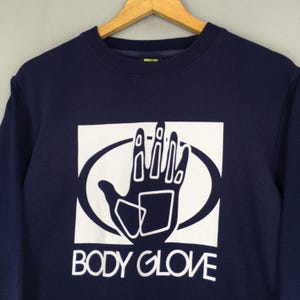 May include: A dark navy blue crewneck sweatshirt featuring a large white graphic print on the chest. The graphic displays a stylised hand silhouette inside an oval shape, with the text BODY GLOVE printed in white capital letters underneath.