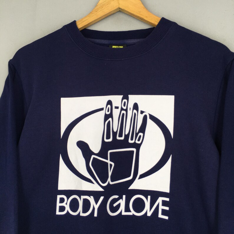 May include: A dark navy blue crewneck sweatshirt featuring a large white graphic print on the chest. The graphic displays a stylised hand silhouette inside an oval shape, with the text BODY GLOVE printed in white capital letters underneath.