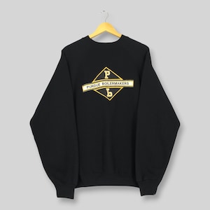 May include: Black Purdue Boilermakers sweatshirt with a gold and white logo. The logo features a diamond shape with the letters "P" and "b" inside, and the words "PURDUE BOILERMAKERS" on a banner. The sweatshirt is hanging on a wooden hanger.