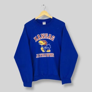 May include: A royal blue sweatshirt with the word "KANSAS" in red and white arched lettering. Below the word "KANSAS" is a cartoon Jayhawk mascot with a yellow beak and blue feathers. The word "JAYHAWKS" is printed below the mascot.