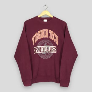 Può includere: Felpa girocollo bordeaux con la scritta "VIRGINIA TECH" in lettere arancioni e bianche. Sotto la scritta c'è la parola "GOBBLERS" in bianco. La felpa è appesa a una gruccia di legno.