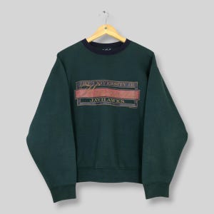 May include: A dark green sweatshirt with a navy blue collar and cuffs. The front features a rectangular design with the words "THE UNIVERSITY OF" above "JAYHAWKS" in a red and gold font. The sweatshirt is hanging on a wooden hanger.