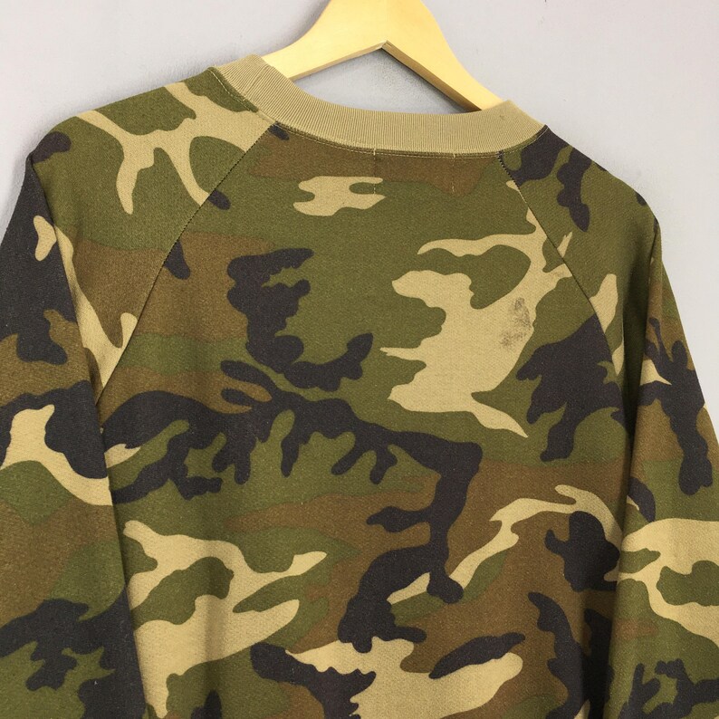 May include: A camouflage-patterned sweatshirt in shades of green, brown, and tan. The long-sleeved top features a crew neck and raglan sleeves. The fabric has a mottled appearance, with the camouflage design covering the entire garment.