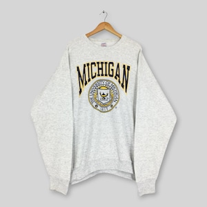 Può includere: Una felpa girocollo grigio chiaro con la scritta MICHIGAN in grandi lettere blu scuro con un bordo giallo. Sotto il testo si trova un sigillo circolare dell'Università del Michigan con una lampada, l'anno 1817 e la scritta THE UNIVERSITY OF MICHIGAN.