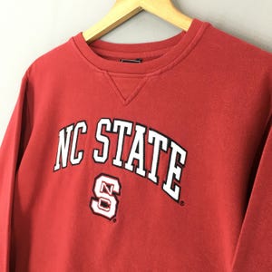 May include: A red long-sleeve crewneck sweatshirt featuring the text NC STATE in white letters with black outlines arched across the chest, positioned above a white and black block letter S logo with a small N inside the center of the letter.