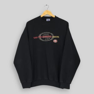 Puede incluir: Sudadera negra de cuello redondo con el logo de los 49ers de San Francisco. El logo presenta el nombre del equipo y un "SF" estilizado en rojo y dorado. Un logotipo más pequeño del equipo está en el lado derecho.