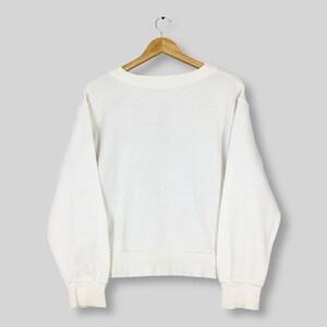 May include: A white crew neck sweatshirt hangs on a wooden hanger. The sweatshirt has long sleeves with ribbed cuffs and a ribbed hem. The fabric appears to be a soft, medium-weight material. The background is a solid, light gray.
