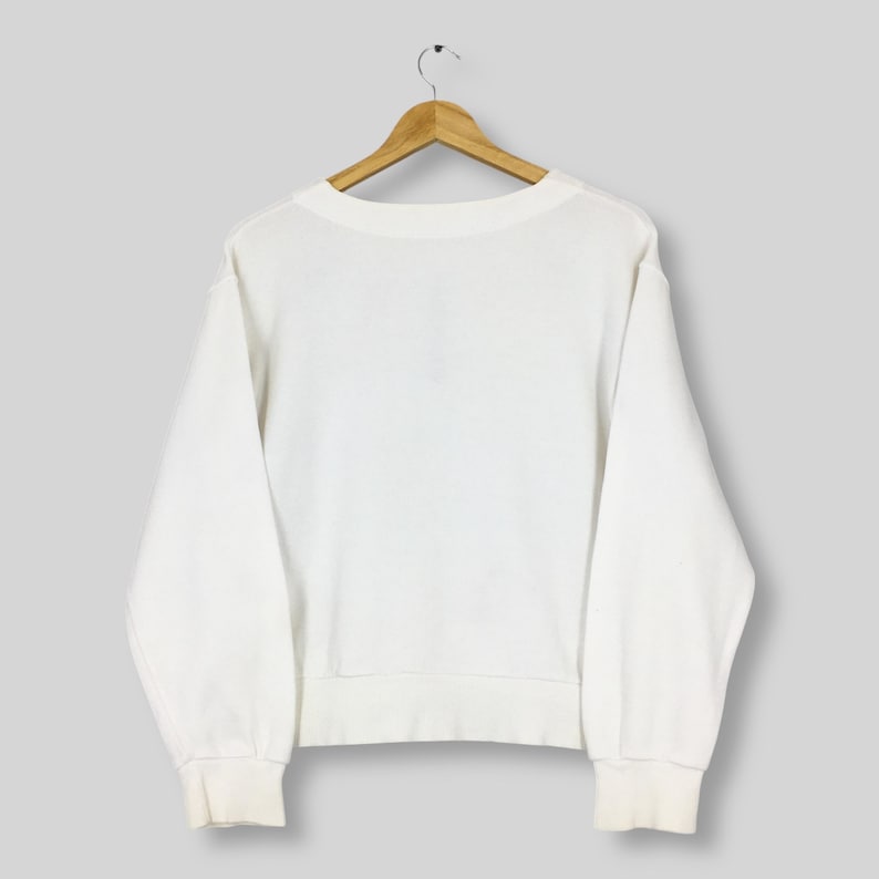 May include: A white crew neck sweatshirt hangs on a wooden hanger. The sweatshirt has long sleeves with ribbed cuffs and a ribbed hem. The fabric appears to be a soft, medium-weight material. The background is a solid, light gray.