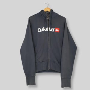 Puede incluir: Una sudadera con capucha con cremallera gris oscuro con el logotipo de Quiksilver en blanco y rojo. La sudadera con capucha tiene una capucha con cordón, una cremallera completa y puños y dobladillo de canalé. La sudadera está colgada en una percha de madera.
