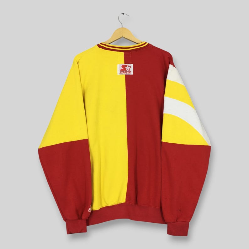 May include: A vintage Starter brand sweatshirt in a color block design. The sweatshirt is primarily yellow and red, with white stripes on the sleeves. The Starter logo is on the chest. The collar has a yellow and red stripe.