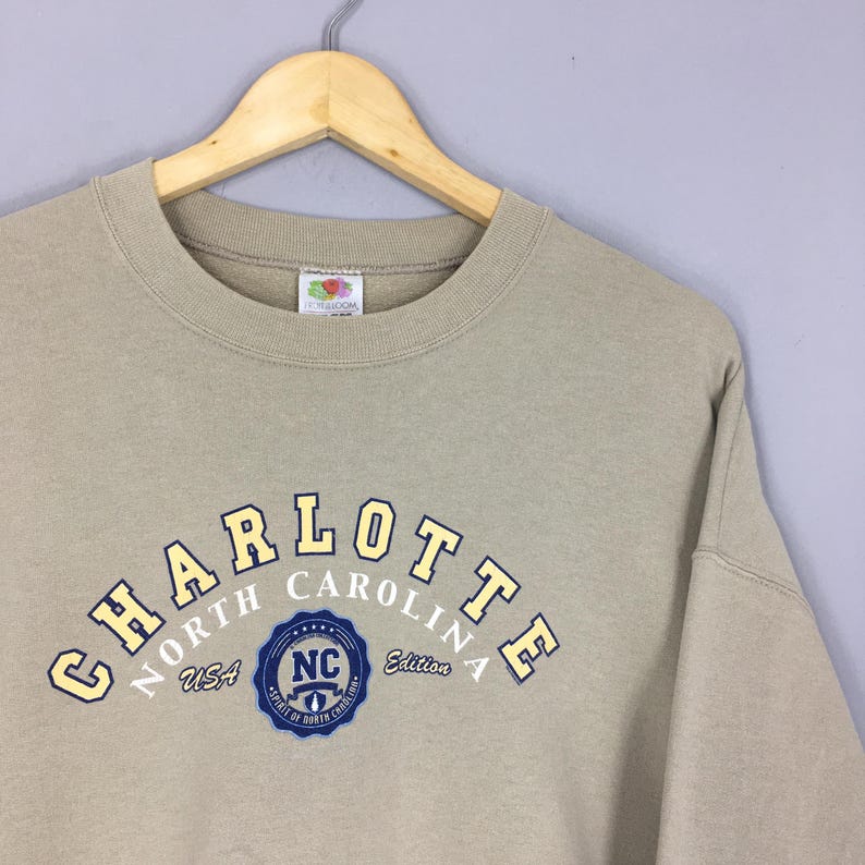 K&ouml;nnte beinhalten: Beiges Sweatshirt mit Rundhalsausschnitt und dem Wort "CHARLOTTE" in gelben und marineblauen Buchstaben auf der Brust. Darunter steht "NORTH CAROLINA USA" und ein blaues Emblem mit "NC".