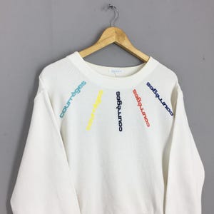May include: White sweatshirt with the word "courr&egrave;ges" embroidered in a rainbow of colors. The text is arranged in a radial pattern across the chest. The sweatshirt is hanging on a wooden hanger.