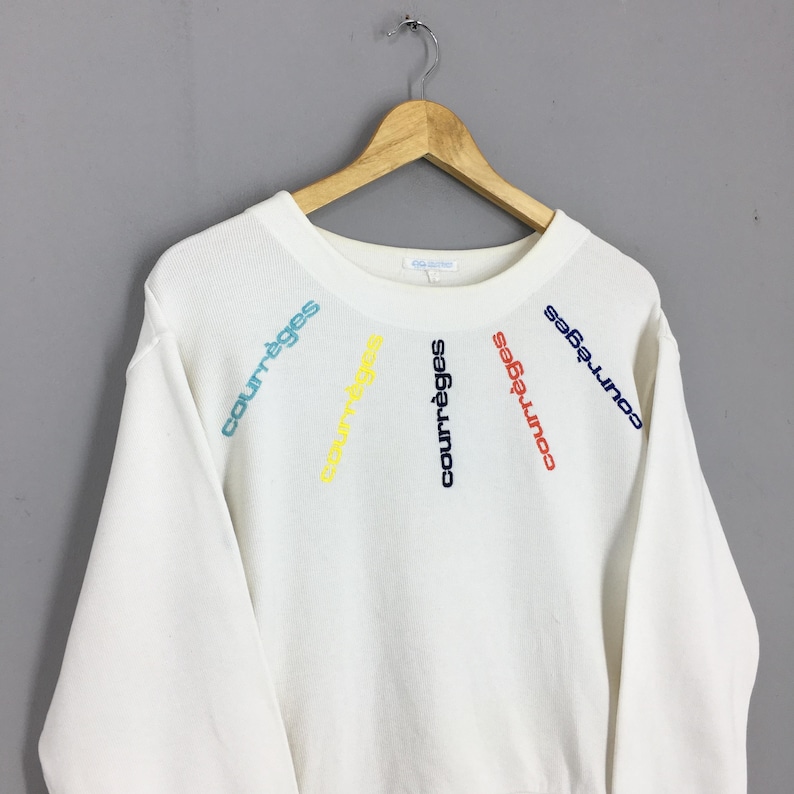 May include: White sweatshirt with the word "courr&egrave;ges" embroidered in a rainbow of colors. The text is arranged in a radial pattern across the chest. The sweatshirt is hanging on a wooden hanger.