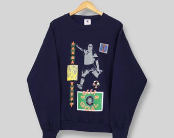 Könnte beinhalten: Marineblaues Sweatshirt mit einem Fußballspieler-Motiv. Das Design umfasst einen Fußball, die Worte "SOCCER GOAL" und geometrische Formen in Gelb, Orange und Grün. Das Sweatshirt hängt an einem Holzkleiderbügel.