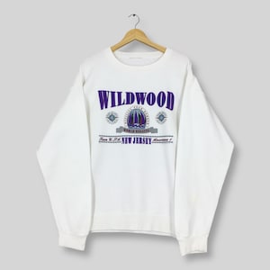 May include: White sweatshirt with the word "WILDWOOD" in purple block letters. Below the word is a graphic of a sailboat and the words "WORLD REGATTA" and "NEW JERSEY". The sweatshirt is hanging on a wooden hanger.