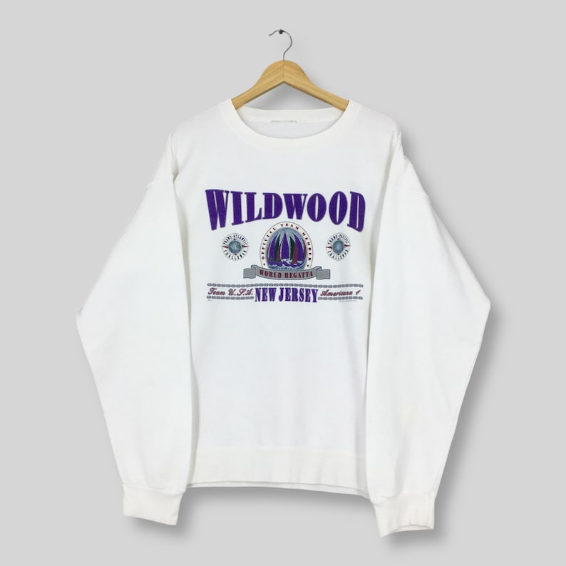 May include: White sweatshirt with the word "WILDWOOD" in purple block letters. Below the word is a graphic of a sailboat and the words "WORLD REGATTA" and "NEW JERSEY". The sweatshirt is hanging on a wooden hanger.