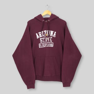 Puede incluir: Sudadera con capucha de color granate con bolsillo delantero y capucha con cordón. El texto blanco en la parte delantera dice "ARIZONA STATE UNIVERSITY" con un gráfico en el centro. La sudadera con capucha está colgada en una percha de madera.