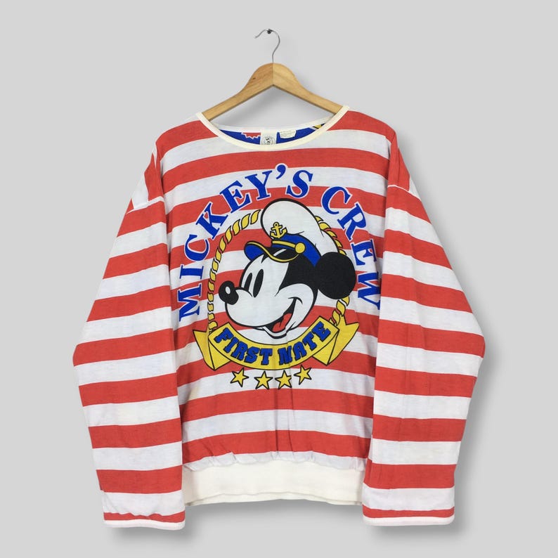 May include: A long-sleeve sweatshirt featuring horizontal red and white stripes. The center graphic displays Mickey Mouse wearing a sailor hat, surrounded by the text Mickey's Crew and First Mate, with a yellow rope border and four yellow stars.