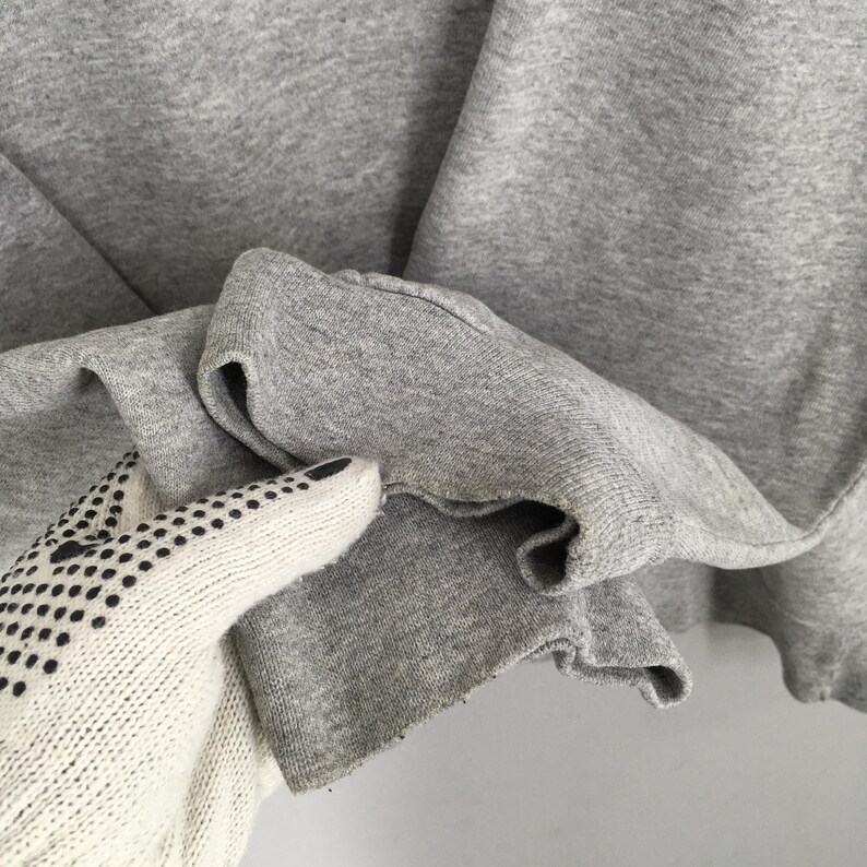 May include: Close-up of a gray sweatshirt sleeve. The cuff is folded over, revealing the inside fabric. The fabric is a solid gray color. A white glove with black dots is touching the sleeve.