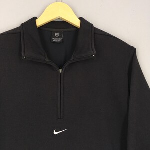 May include: Black athletic top with a white Nike logo. The top features a ribbed texture, a collar, and a half-zip. The top is suspended on a wooden hanger. The garment is made of a dark material.
