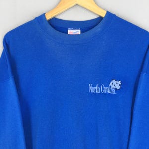 May include: A blue sweatshirt with the words "North Carolina" and a logo printed on the front. The sweatshirt is made of a soft material and has a crew neck. The garment is hanging on a wooden hanger.