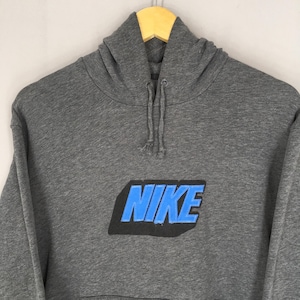 Może przedstawiać: Szara bluza z kapturem z napisem "NIKE" w niebieskich literach na przodzie. Bluza ma kaptur ze sznurkiem i kieszeń typu kangur. Odzież wisi na drewnianym wieszaku.