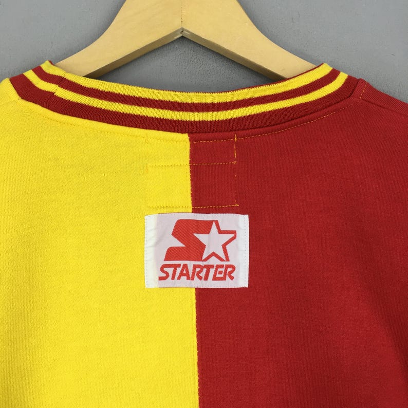 May include: A vintage crew neck sweatshirt, split vertically with yellow and red panels. The collar is striped with red and gold. A white rectangular patch with the "Starter" logo is centered on the lower back.