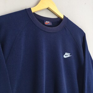 May include: A navy blue Nike sweatshirt with a crew neck and long sleeves. The Nike logo is embroidered in white on the chest and on the label inside the collar. The sweatshirt is hanging on a wooden hanger.