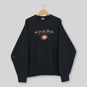 Könnte beinhalten: Ein schwarzes Langarm-Sweatshirt mit Rundhalsausschnitt, das auf der Brust den in Gelb und Rot gestickten Schriftzug 49ERS SAN FRANCISCO sowie ein kleines ovales 49ers-Teamlogo direkt unter dem Text auf der Vorderseite des Kleidungsstücks aufweist.