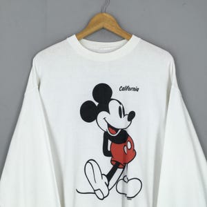 May include: White long-sleeve shirt featuring a smiling Mickey Mouse graphic in black, red, and white. The word "California" is printed above the character. The shirt is hanging on a wooden hanger.