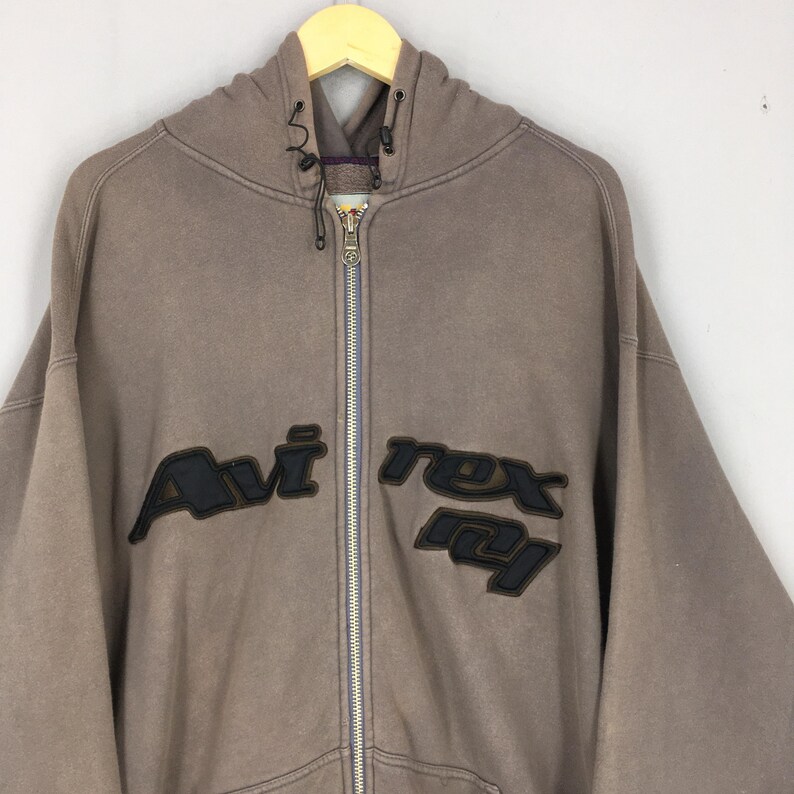 Puede incluir: Una sudadera con capucha marr&oacute;n con cremallera y el texto "Avi Rex" en letras negras en el pecho. La sudadera con capucha tiene una capucha con cord&oacute;n y una cremallera completa. La tela parece ser un material suave y desgastado.